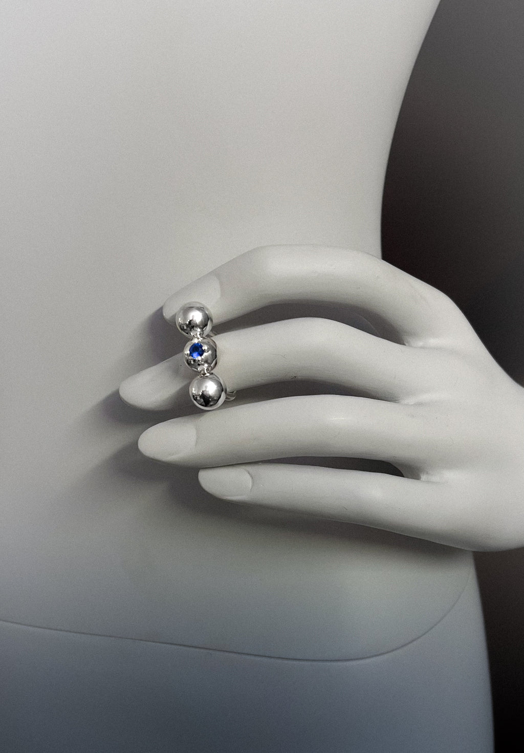 Silver ring made out of spheres with a blue gemstone on a mannequin hand against a neutral background