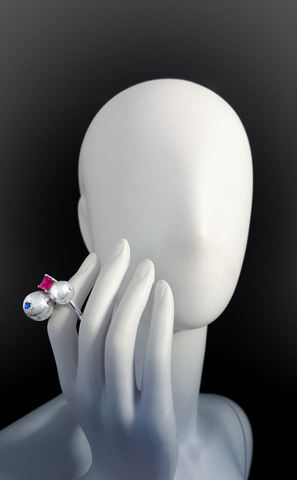 Mannequin hand wearing a ring with a pink and blue gemstone on a black background. statement ring